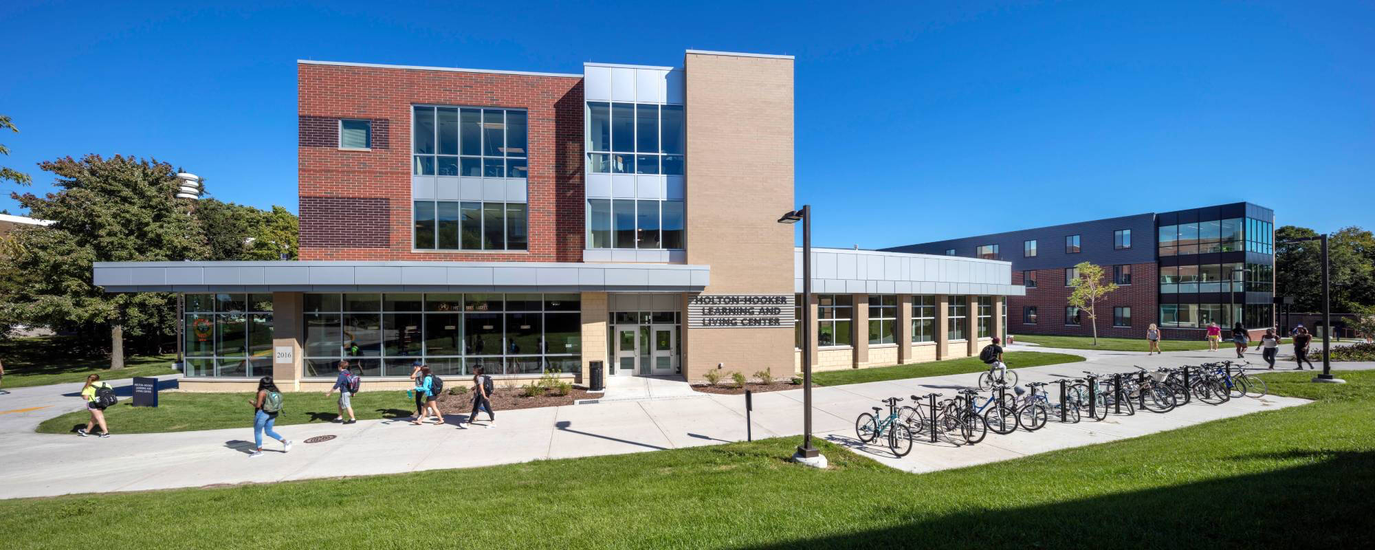Modern two-story academic building, Holton-Hooker Learning and Living Center, with large windows and a mix of brick and beige exterior. Bicycles are parked outside, and people walk on paths under a clear blue sky.